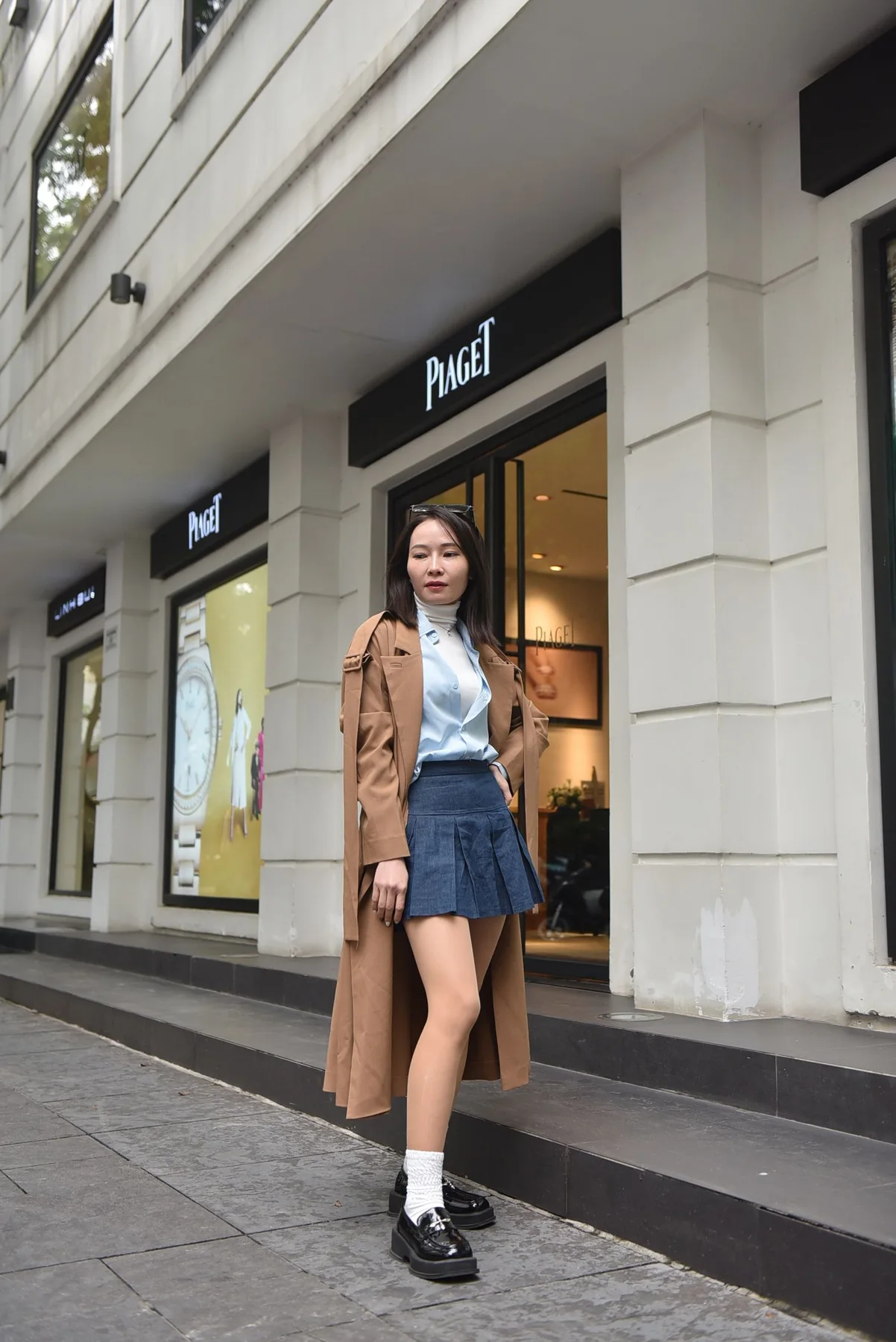A woman in a long brown outer layer and short pleated skirt on a city pavement.