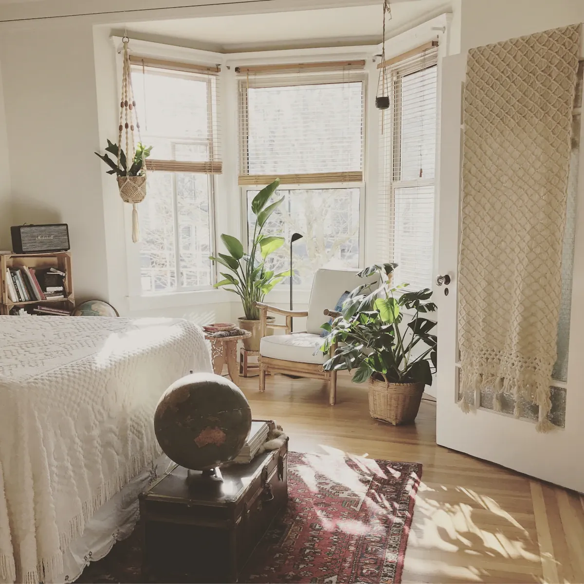 A pale upholstered chair beside bay windows and plants in a bedroom.