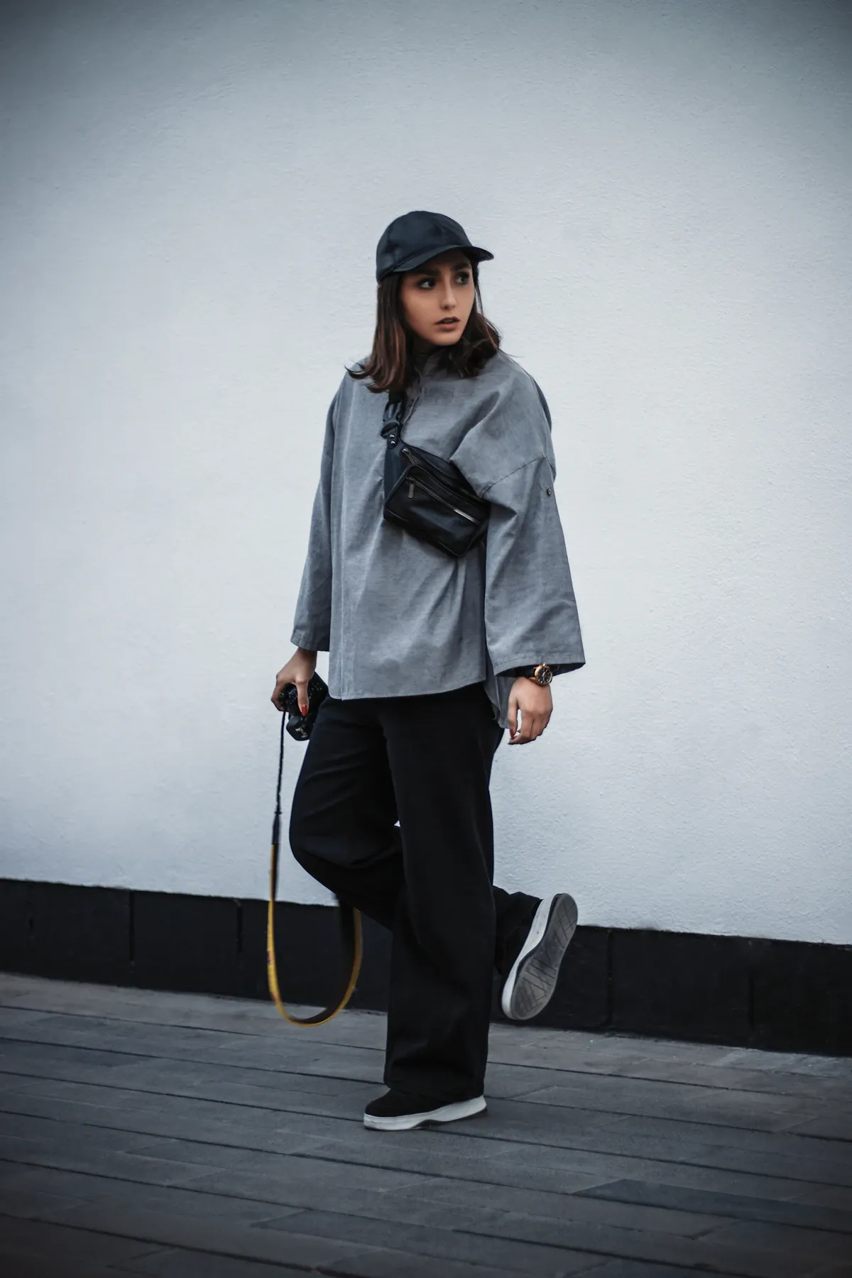 A woman wearing a grey shirt, dark trousers and a dark cap beside a pale wall.
