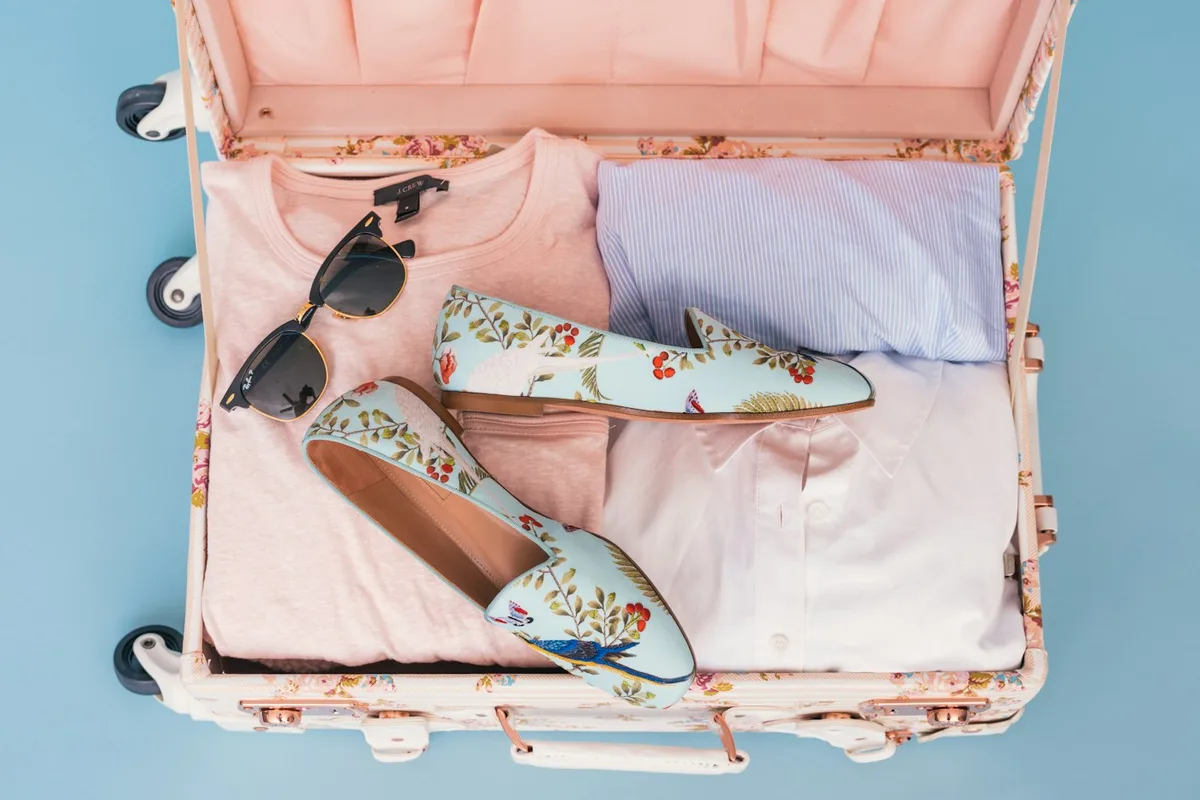 An open pale pink suitcase with clothing, floral shoes and sunglasses.