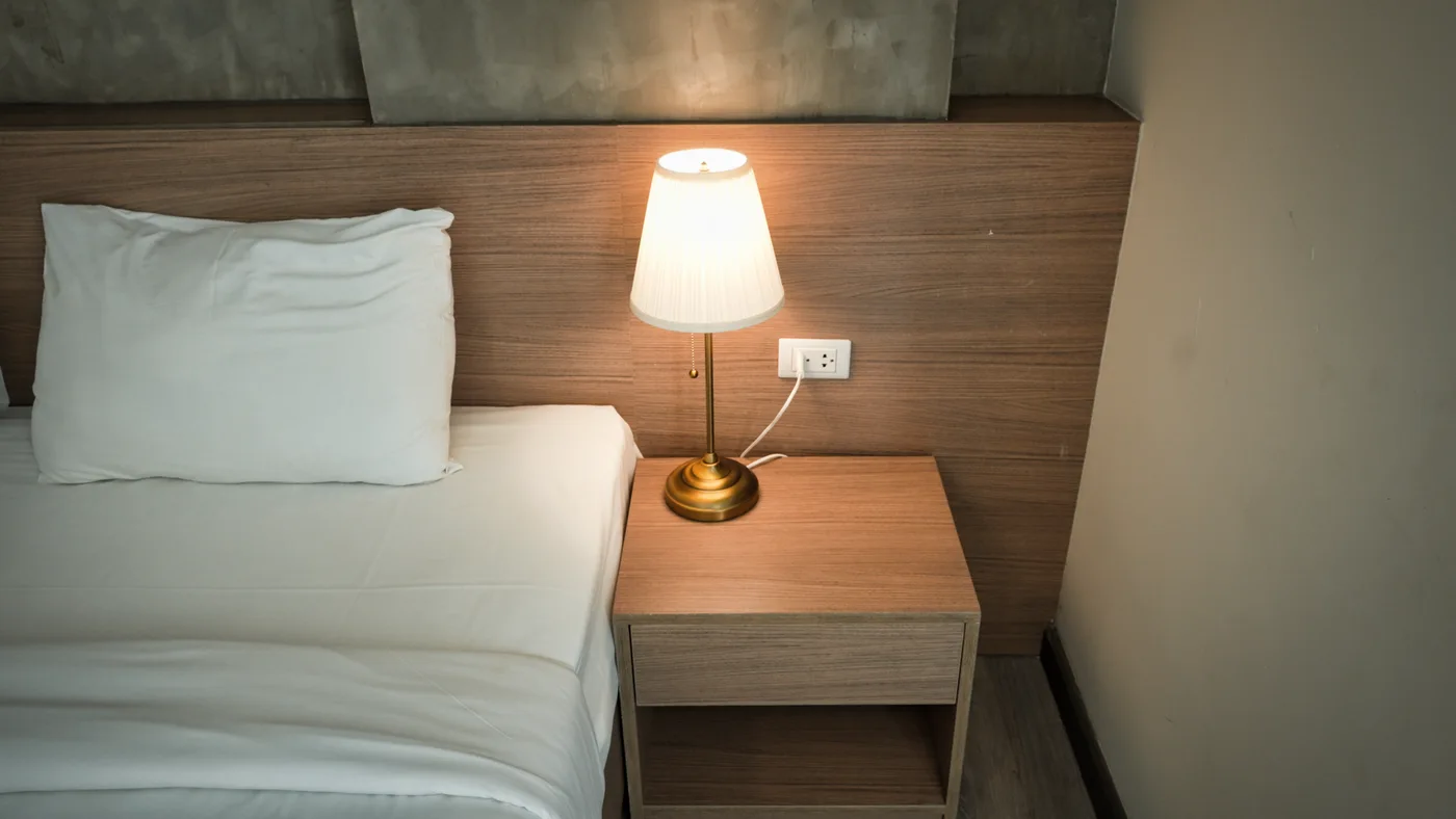 A lit bedside lamp on a wooden table beside pale bedding and a wooden headboard.