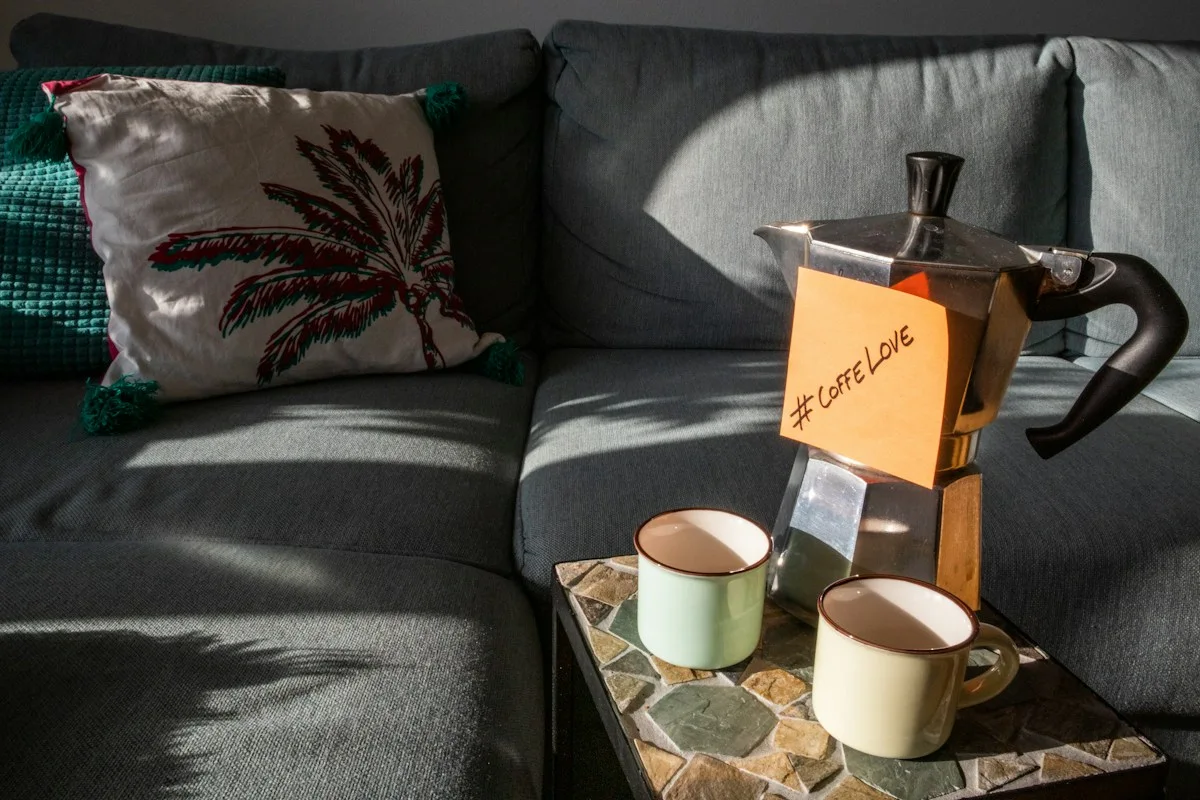 Two cups and a coffee pot beside a sofa with a patterned cushion.