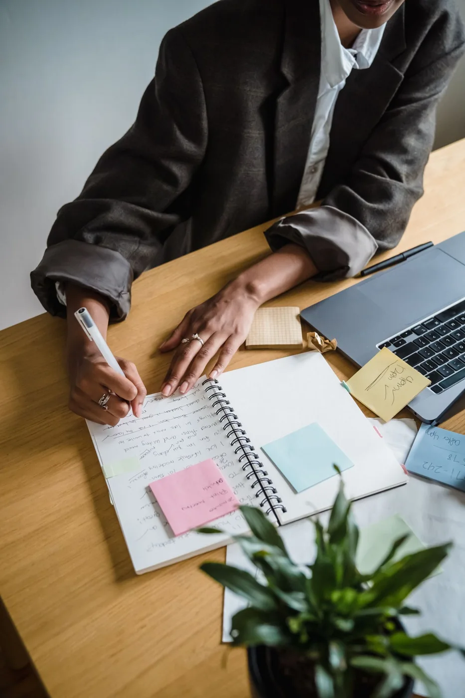 A person writing at a desk with a notebook, papers and a laptop.