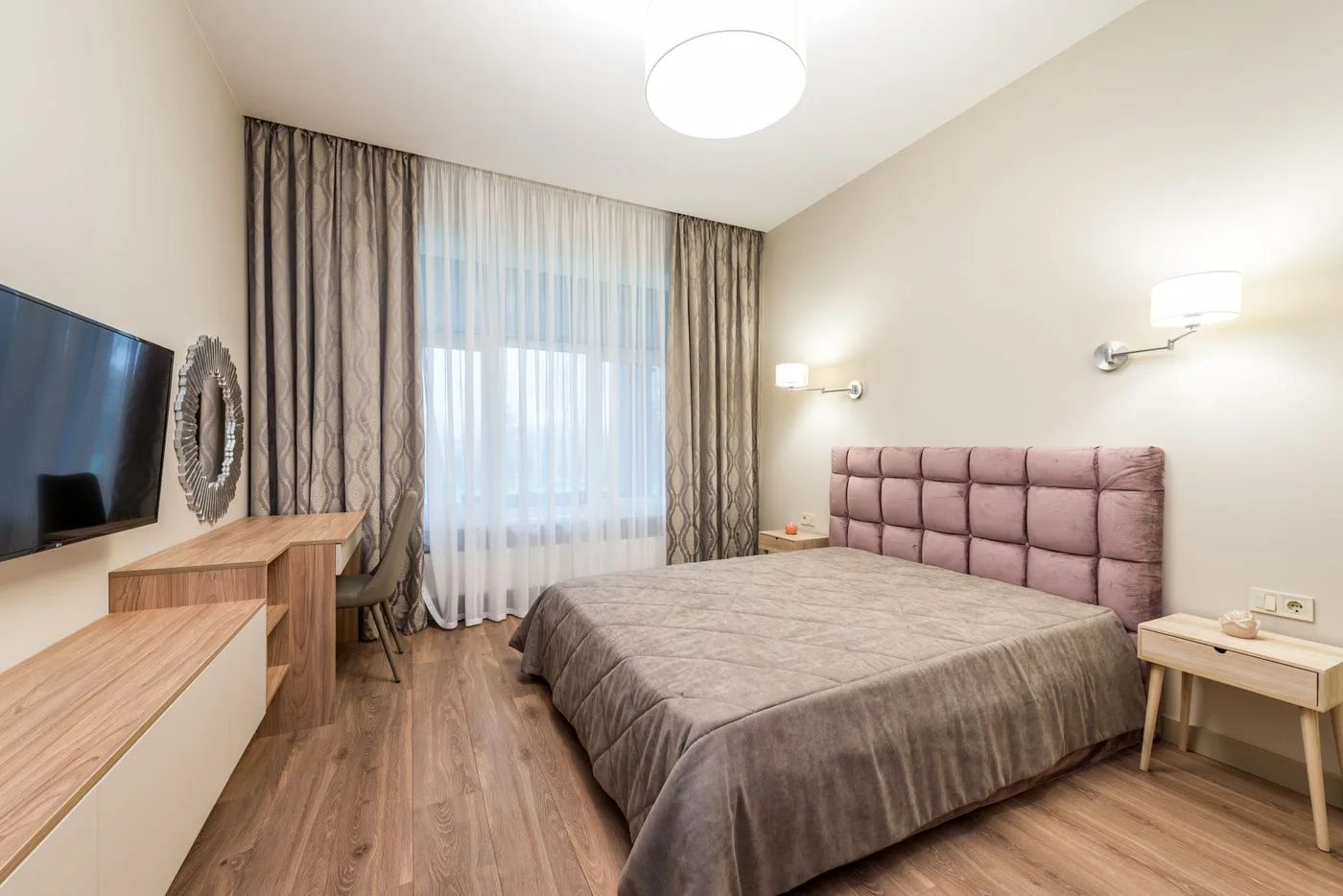 A bedroom with pale curtains, a television and a pink upholstered headboard.