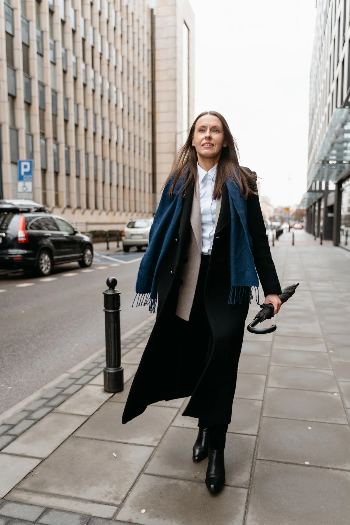 A woman in a long dark coat holding a folded black umbrella on a city street.