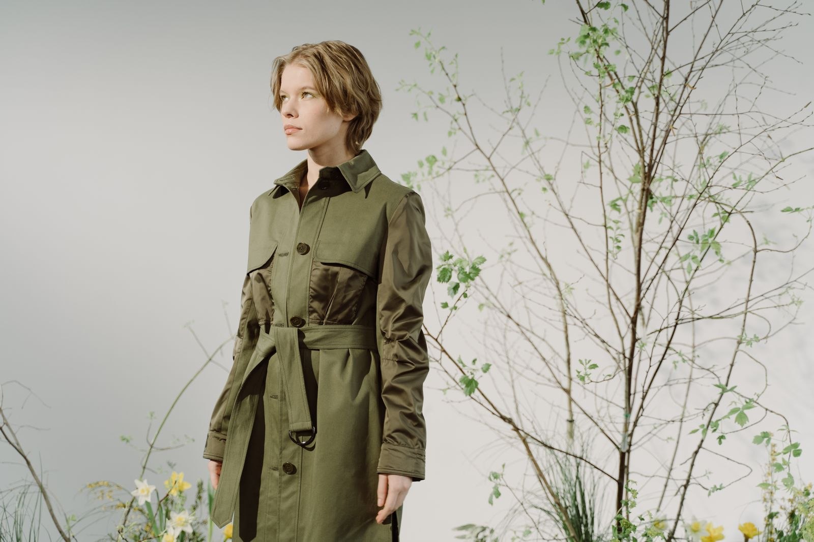 A woman wearing a belted green outer layer beside plants.