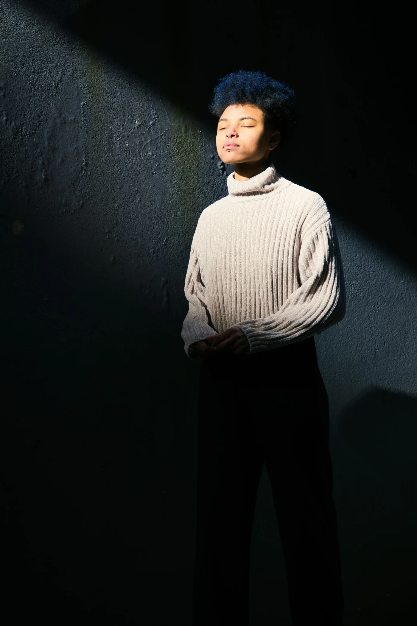 A person wearing a pale high-necked top beside a sunlit wall.